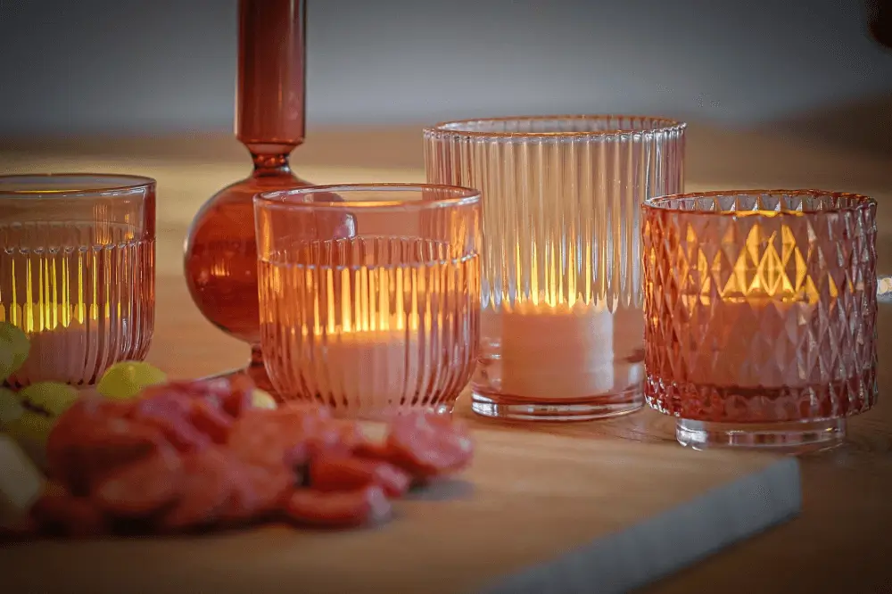 Vier verlichte led waxinelichtjes in roze glazen houders met textuur zitten op een houten oppervlak naast een schotel met gesneden vleeswaren en kaas en creëren een warme en gezellige sfeer.