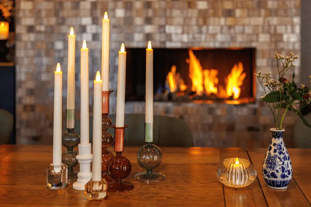 Een groep brandende kaarsen in glazen houders staat op een houten tafel, naast led waxinelichtjes, een kleine blauw-witte vaas met bloemen en een waxinelichtje. Op de achtergrond zorgt een open haard met vlammen voor een warme, gezellige sfeer.